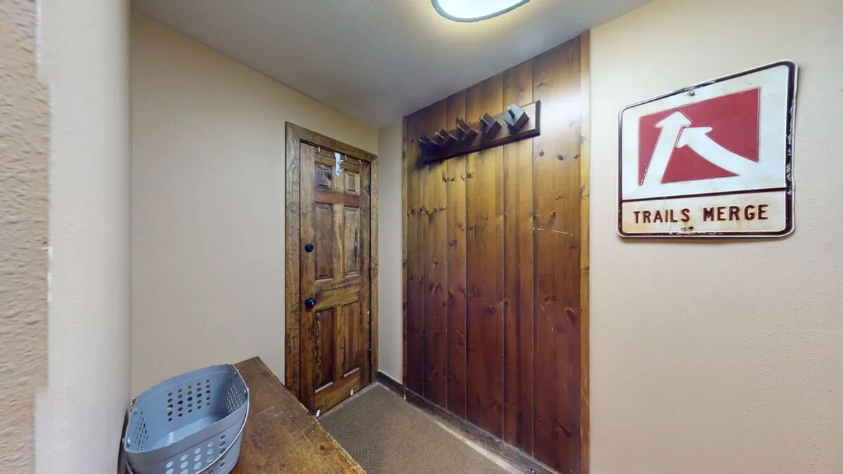 This image captures a rustic mudroom or entry area featuring a 'TRAILS MERGE' sign, wall hooks for gear, a wooden bench, and a sturdy wood door. It embodies the transition point between outdoor activity and indoor comfort.