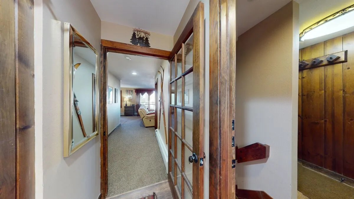 The image captures the entrance door opening up into a hallway and living area, featuring a warm wood wall, a mirror, and inviting furnishings. The scene is cozy, yet it hints at movement and flow—people coming and going, gathering after a day outside.