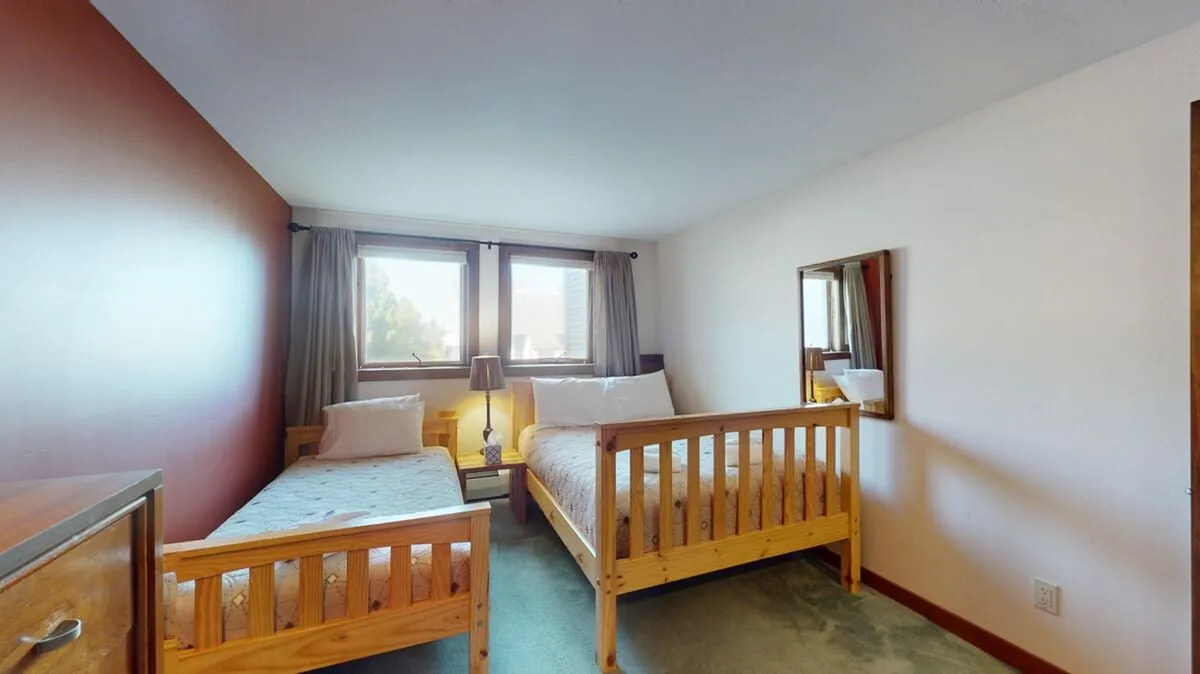 A shared bedroom with two wooden beds, large windows letting in natural light, and a nightstand with a lamp. The inviting atmosphere is ideal for sharing stories before bed or enjoying morning sun after a restful night.