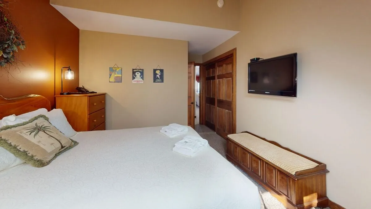 A serene master bedroom featuring a plush bed adorned with soft linens, a wall-mounted flat-screen television, tasteful wall art, and a storage bench at the foot of the bed. The room is suffused with natural light, creating a restful and inviting atmosphere.