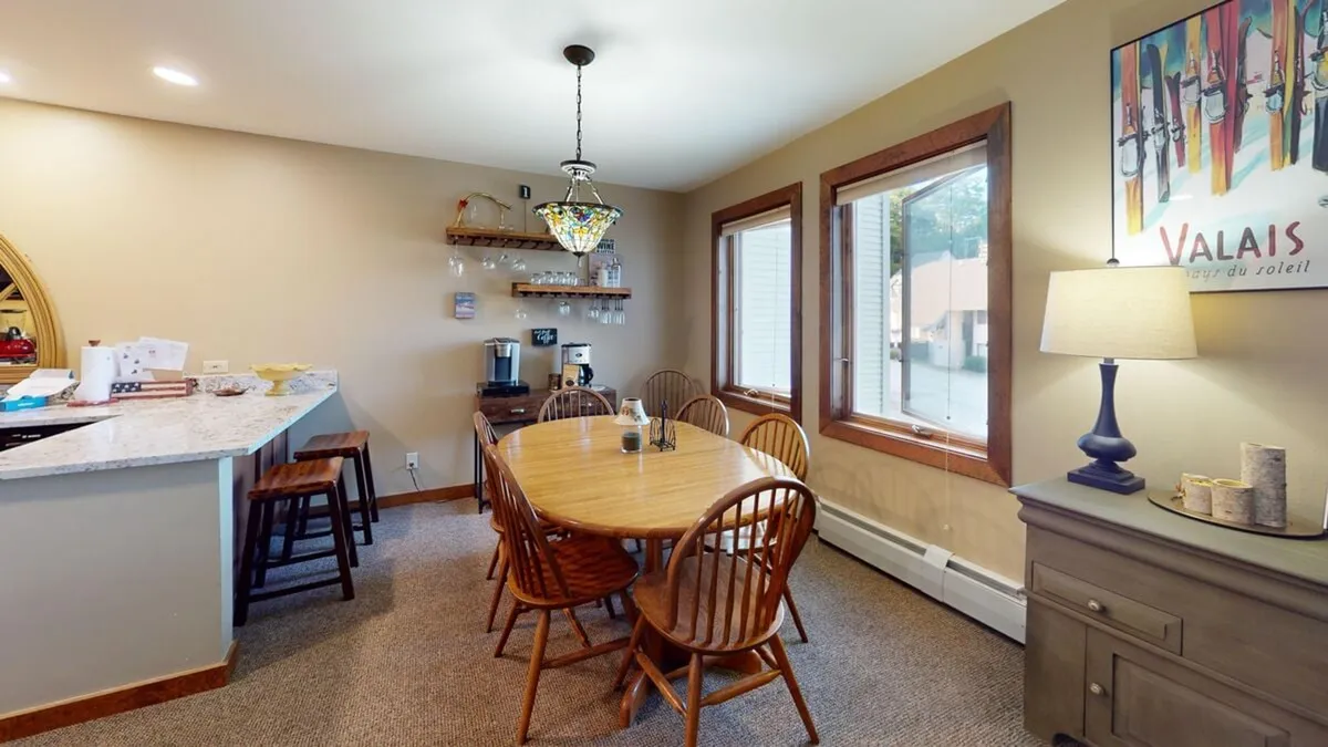 An open dining area centered around an elegant oval table for eight, illuminated by a stylish chandelier overhead. Bar stools along the kitchen counter and decorative art add to the convivial, upscale ambiance, suggesting a space perfect for group meals and celebrations.