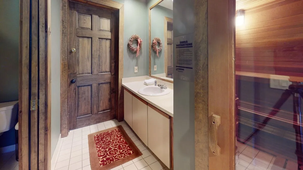 A bathroom featuring a vanity with basin, mirror, wooden door, and cozy decorative elements like wall wreaths and a rug. The space exudes tranquility and is perfect for preparing to enjoy the in-house sauna or for unwinding after a day outdoors.