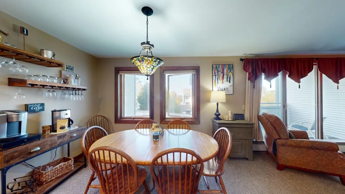 A cozy, welcoming dining space featuring a large table, a colorful chandelier, and a wine glass rack, with ample windows bringing in natural light. The scene is set for family meals, intimate gatherings, or enjoying a glass of wine after a day outdoors.